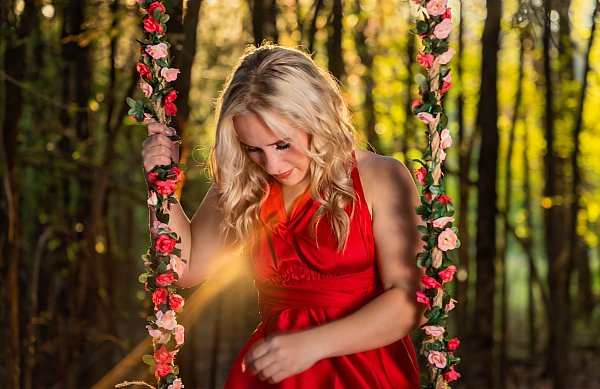 Senior portrait photographer North Little Rock Arkansas — high school senior in red dress on flower-covered swing in golden hour forest light, Liza Jane Photography